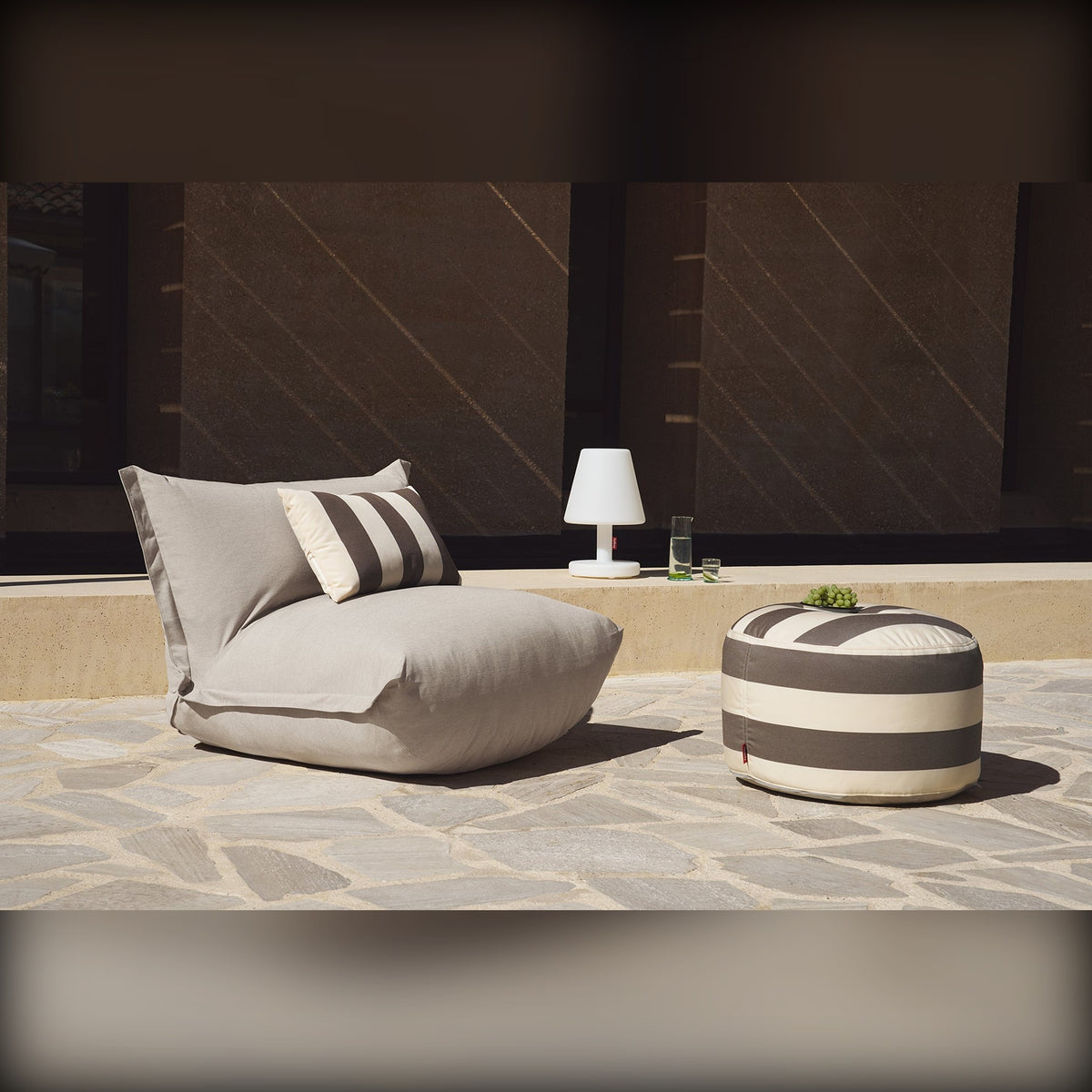 Bonbaron Outdoor Grey Taupe Domestica – Lifestyle – On a Stone Patio with Striped Cushion, Pouf and Table Lamp